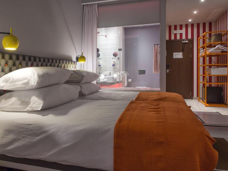 Modernes Hotelzimmer mit großem Bett, weißen Wänden, orangen Bettdecke und gläsernem Badezimmer.