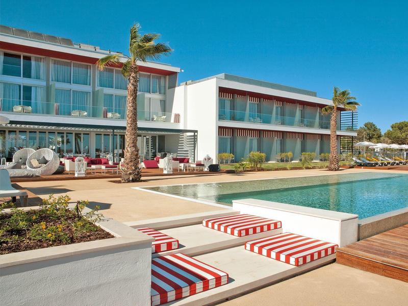 Modernes Hotel mit Pool, Palmen und roten weißen Liegestühlen unter klarem blauen Himmel.