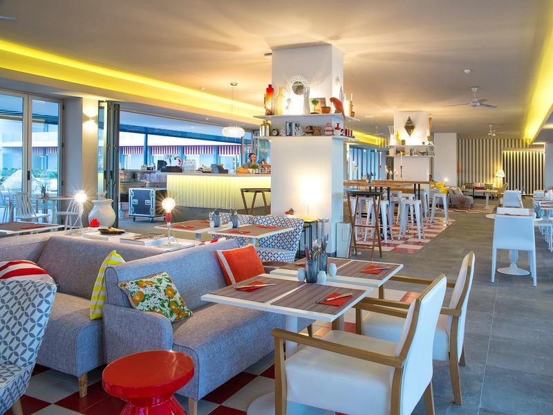 Moderner Gastraum mit grauen Sofas, bunten Kissen, Holzstühlen und Bar mit Beleuchtung.