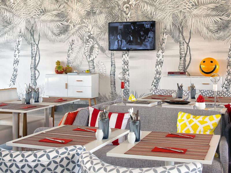 Hell eingerichteter Gastraum mit Holztischen, roten Tischsets, gemusterten Kissen und Wandbild mit Pflanzenmotiven.