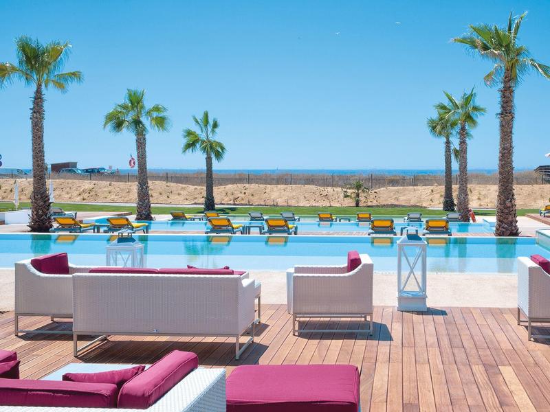 Poolbereich mit weißen Sofas, pinken Kissen, Holzterrasse und Palmen am Meer im Hintergrund.