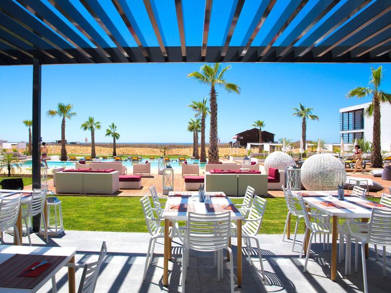 Terrassenbereich mit Tischen, Stühlen, Palmen und blauem Himmel am Meer im Hintergrund.