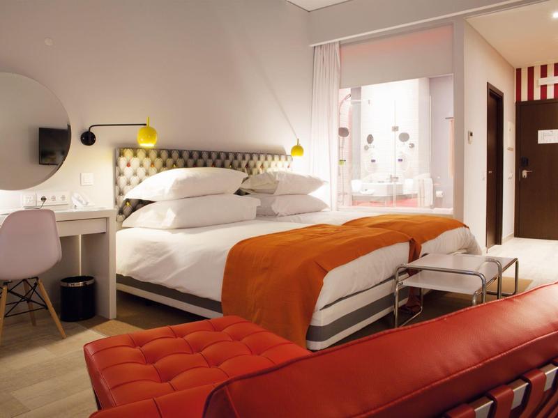 Modernes Schlafzimmer mit großem Bett, orangefarbener Decke und rotem Ledersofa im Vordergrund.