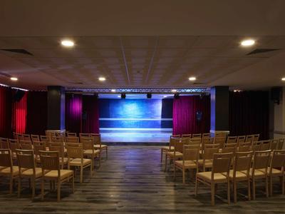 Leerer Theatersaal mit Holzbühne, roten Vorhängen und Holzstühlen in Reihen angeordnet.