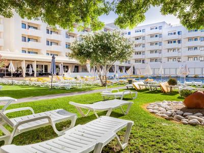 Grüner Garten mit weißen Liegestühlen vor einem mehrstöckigen Hotelgebäude unter Bäumen.