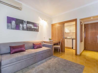 Kleines Wohnzimmer mit grauer Couch, lila Kissen, Teppich und offener Küchenzeile in warmem Licht.