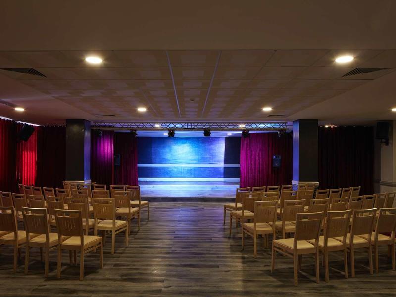 Leerer Theatersaal mit Holzbühne, roten Vorhängen und Holzstühlen in Reihen angeordnet.