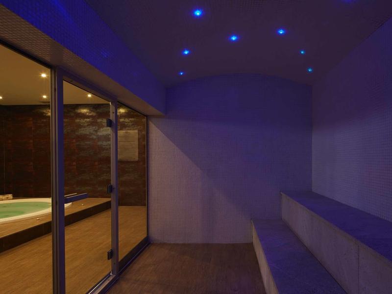 Innenraum mit blauer Beleuchtung, Glaswand und Holzboden mit breiter Sitzbank aus Holz.