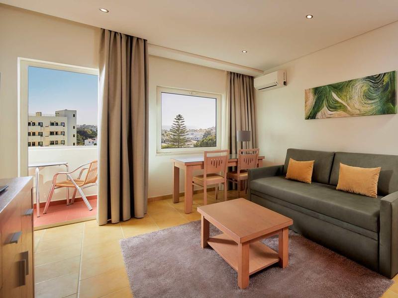 Helles Wohnzimmer mit Sofa, Tisch, Teppich, Fenster, Balkon und moderner Einrichtung in warmen Farbtönen.