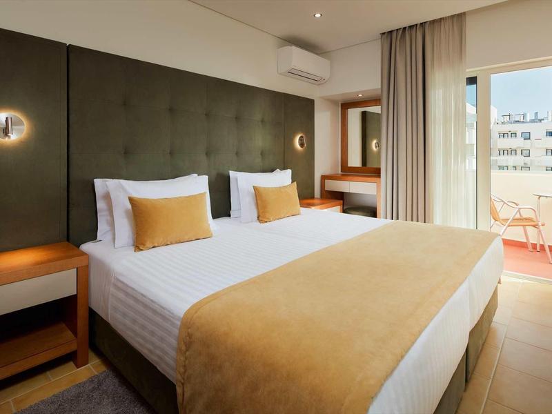 Modernes Schlafzimmer mit großem Bett, hellen Wänden, grünem Kopfteil und Balkon mit Stadtblick.