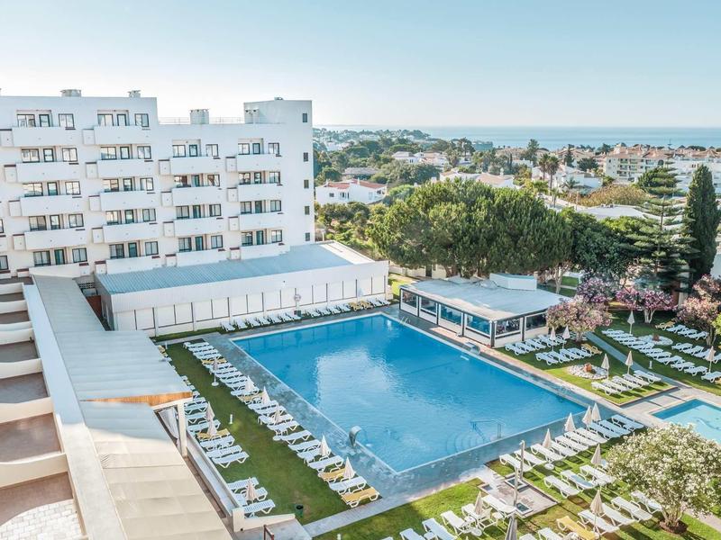 Großer rechteckiger Pool im Freien mit weißen Liegestühlen, umgeben von mehrstöckigem Gebäude und Bäumen.