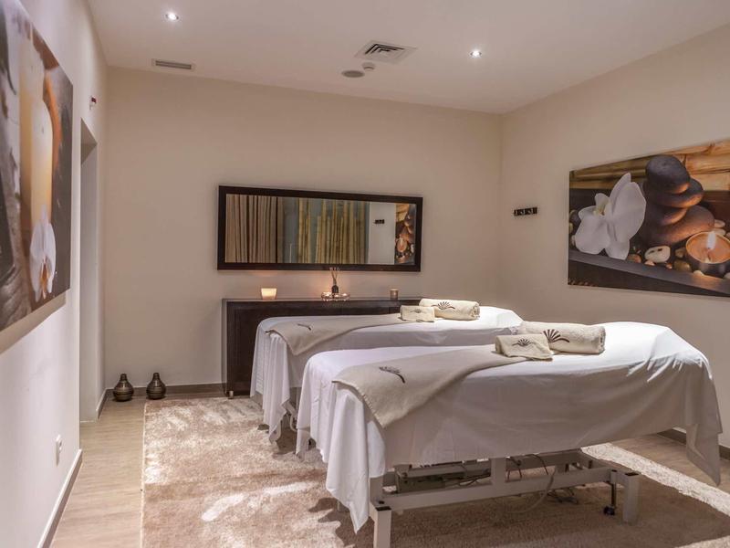 Heller Massageraum mit zwei weißen Liegen, beruhigender Kunst an den Wänden und warmem Licht.