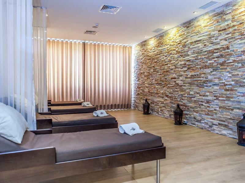 Entspannungsraum mit Liegen, Steinwand, gedämpftem Licht und großen Fenstern mit Vorhängen.