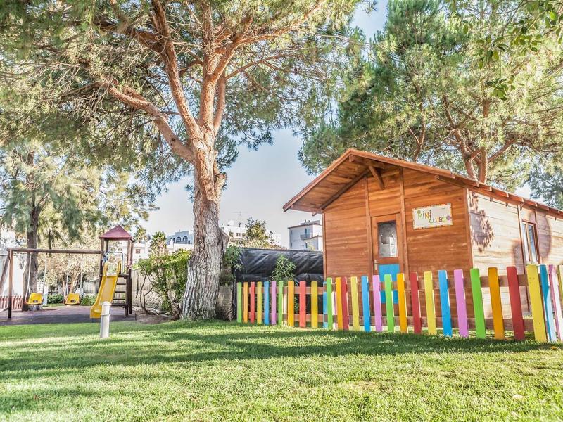 Ein bunter Gartenzaun umgibt ein kleines Holzhaus im grünen Garten unter großen Bäumen.