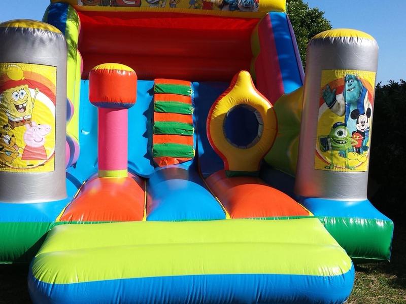 Bunte Hüpfburg mit Comics, gelb-blau-rot-grün, auf Gras mit blauem Himmel im Hintergrund.