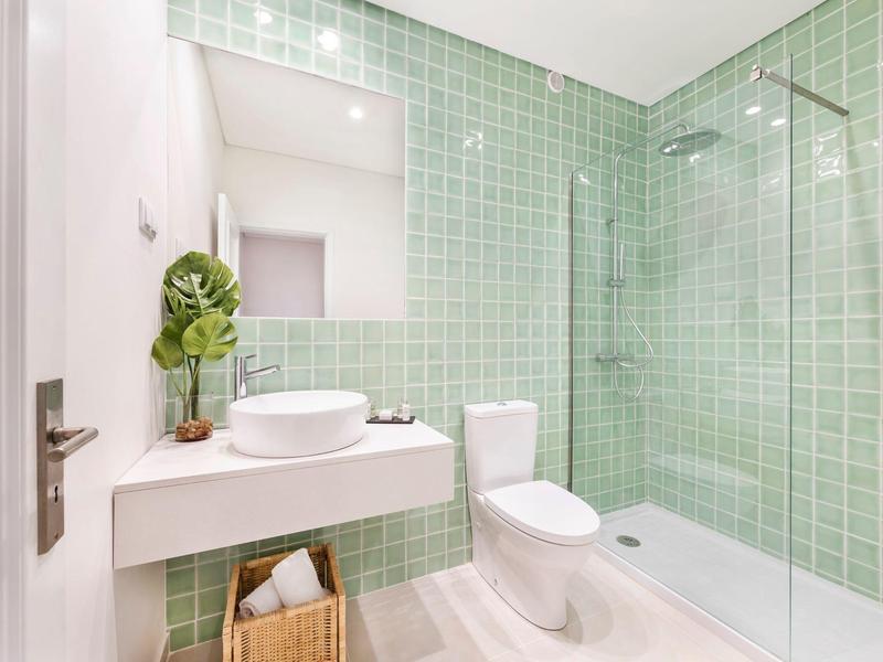 Modernes Badezimmer mit grün gekachelter Dusche, weißem WC, ovalem Waschbecken und Holzkorb.