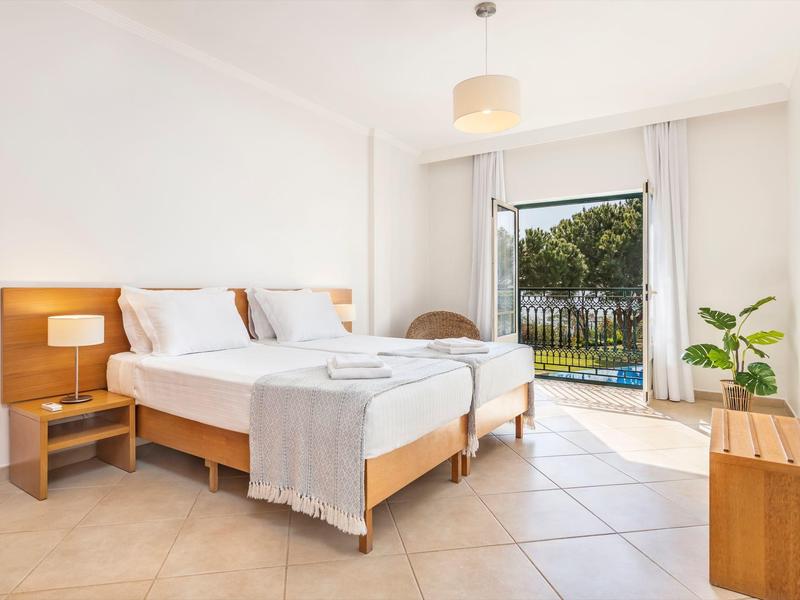 Hell eingerichtetes Schlafzimmer mit zwei Betten, Nachttisch, Sitzstuhl und Terrasse mit Blick ins Grüne.