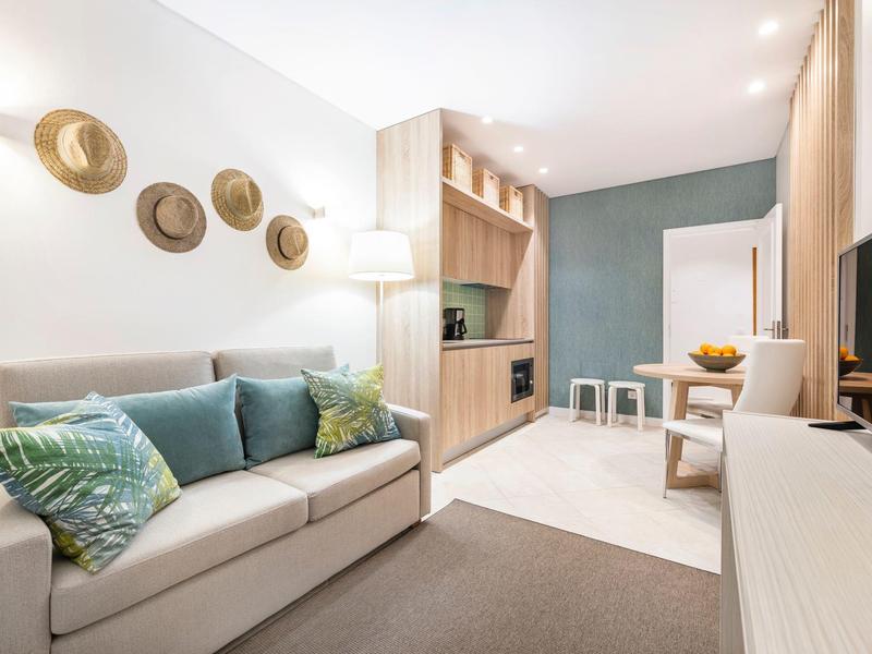 Modernes Wohnzimmer mit beigem Sofa, grünen Kissen, Holzregalen und Strohhalm-Hut-Dekor an der Wand.