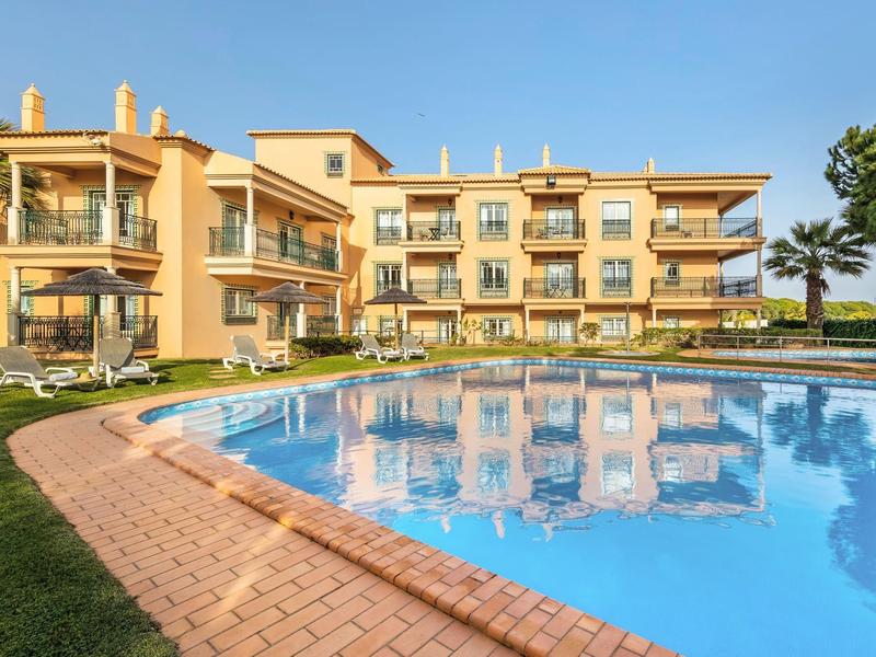 Großes, gelbliches Hotelgebäude neben rundem Pool mit blauer Wasseroberfläche und grüner Wiese.