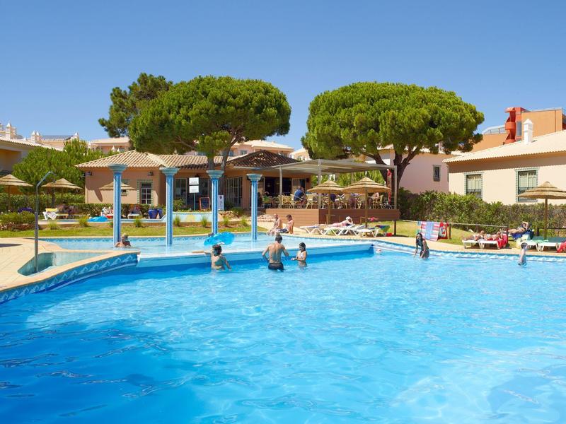 Großer blauer Pool mit Menschen, Sonnenschirmen und grünen Bäumen bei sonnigem Himmel.