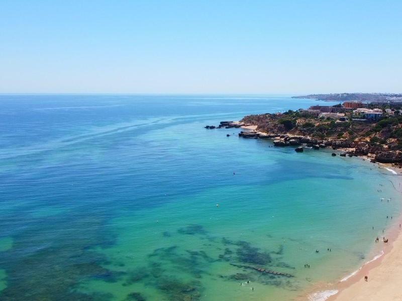 Klarer blauer Himmel über ruhigem, türkisfarbenem Meer und sandigem Strand mit Felsen im Hintergrund.