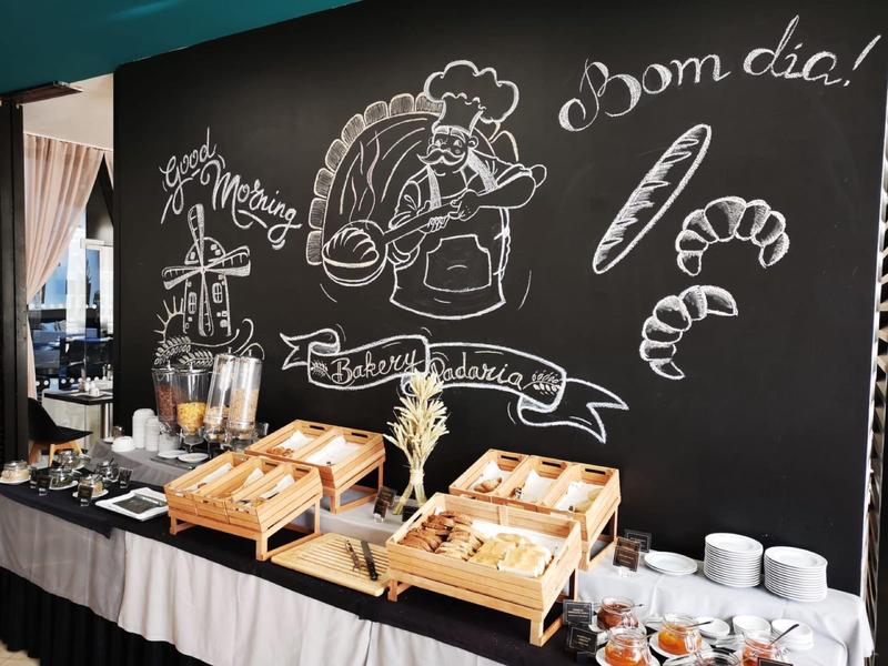 Buffet mit Backwaren, Croissants, Getränkespendern und Tellern vor großer schwarzer Wandtafel.