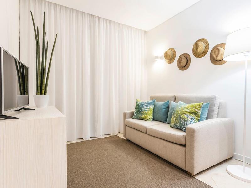 Kleines Wohnzimmer mit grauem Sofa, bunten Kissen, Wanddeko aus Hüten und Stehlampe.