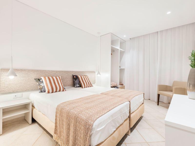 Helles Schlafzimmer mit zwei Betten, beige Bettwäsche, weißen Wänden und einem kleinen Tisch mit Stuhl.