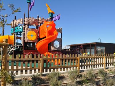 Buntes Piratenschiff-Spielgerüst mit Rutsche auf Spielplatz, umgeben von Holzzaun und Pflanzen.