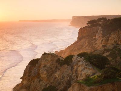 Küste mit steilen Felsen und Bäumen im warmen Licht des Sonnenuntergangs über ruhigem Meer.