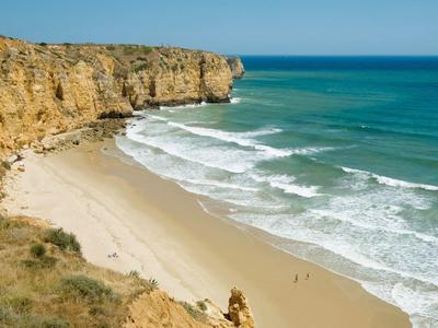 Ein weiter Sandstrand mit Wellen, umgeben von hohen, gelblichen Felsen und blauem Himmel.