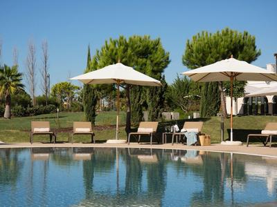 Schwimmbecken mit Sonnenschirmen, Liegestühlen und grünen Bäumen vor klarem blauem Himmel.
