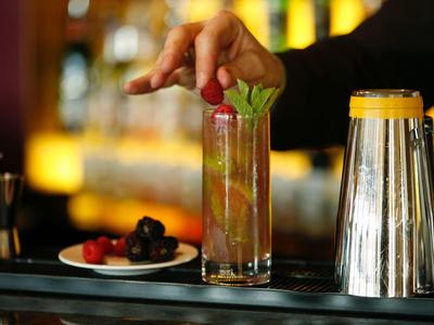 Hand legt Minzblatt in ein hohes Glas mit Cocktail, rote Beeren und Strohhalm, Barhintergrund.