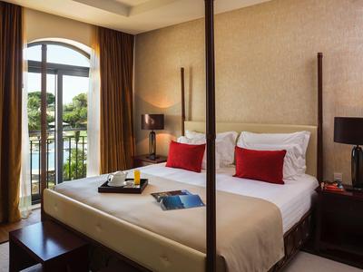 Helles Hotelzimmer mit Doppelbett, roten Kissen und Blick auf einen Balkon mit Wasserblick.