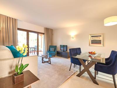 Helles Wohnzimmer mit blauen Sesseln, Sofa, Holztisch, Teppich und großem Fenster mit Vorhängen.