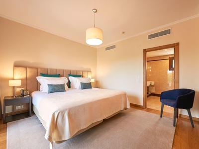 Helles Schlafzimmer mit Doppelbett, Nachttischen, blauer Sitzgelegenheit und offenem Badezimmertür.