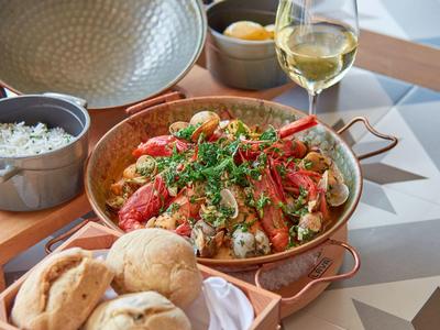 Tabakpfanne mit Hummer, Reis, Brot und Weißwein auf gemusterter Tischoberfläche serviert.