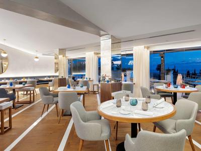 Moderner, heller Restaurantraum mit weißen Tischen, grauen Stühlen und großen Fenstern mit Ausblick.