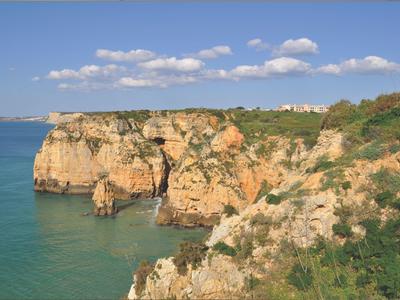 Klippen mit orange-braunem Felsen an ruhigem türkisfarbenem Meer unter blauem Himmel mit Wolken.