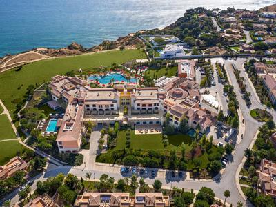 Großer Hotelkomplex mit Pool, Grünflächen und Meer im Hintergrund, umgeben von Straßen und Häusern.
