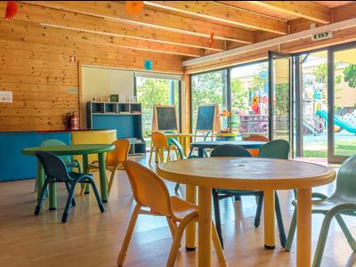 Bunter Kindergartenraum mit Holzwänden, großen Fenstern und verschiedenen Tischen und Stühlen.