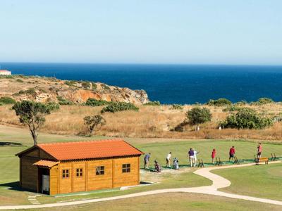 Grüne Landschaft mit kleinem Holzhäuschen und Menschen vor einer Küste und blauem Meer.