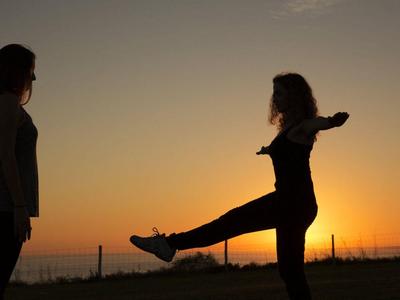 Zwei Frauen bei Sonnenuntergang, eine steht, die andere macht einen Bein-Kick im Freien.