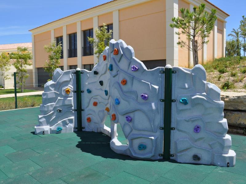 Outdoor-Kletterwand mit bunten Griffen vor einem modernen gelben Gebäude und grünem Bodenbelag.