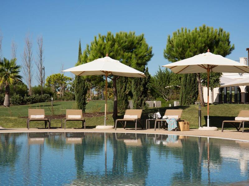 Schwimmbecken mit Sonnenschirmen, Liegestühlen und grünen Bäumen vor klarem blauem Himmel.