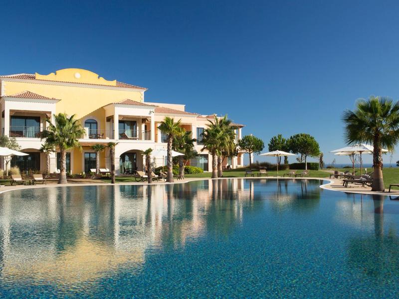 Großer, blauer Pool vor gelblichem Hotelgebäude mit Palmen und einem klaren Himmel im Hintergrund.