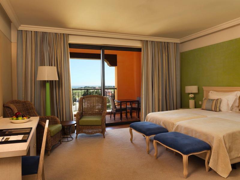 Helles Hotelzimmer mit Doppelbett, blauen Hockern, Schreibtisch, Sessel und Balkon mit Blick ins Freie.