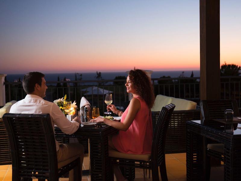 Paar sitzt bei Sonnenuntergang draußen, trinkt Wein und genießt romantisches Abendessen.