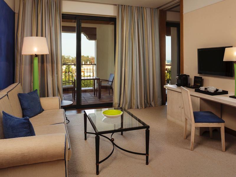 Gemütliches Wohnzimmer mit Sofa, Glastisch, Schreibtisch und Zugang zu Balkon mit Vorhängen.