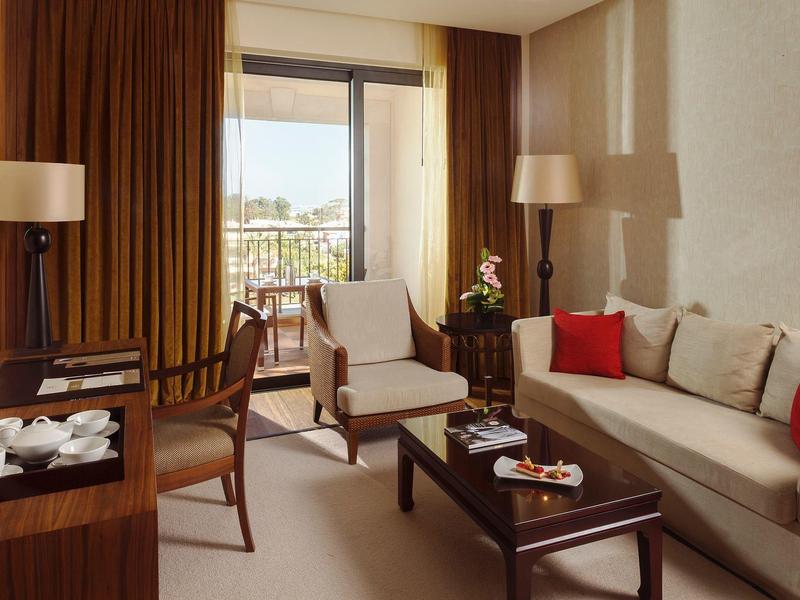 Wohnzimmer mit beige Sofa, roten Kissen, zwei Sesseln, Holztisch und großem Fenster mit Blick.